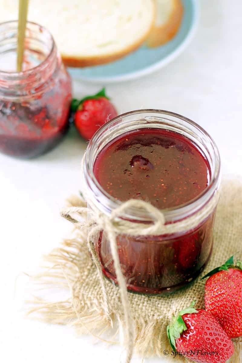 a small mason jar filled with strawberry jam made in the instant pot in the centre and a white bread ready to be smeared with jam in the background