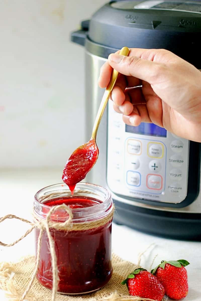 my fingers are holding a spoon with strawberry jam made in the instant pot which is dripping into the mason jar showing the texture with an instant pot int he background.