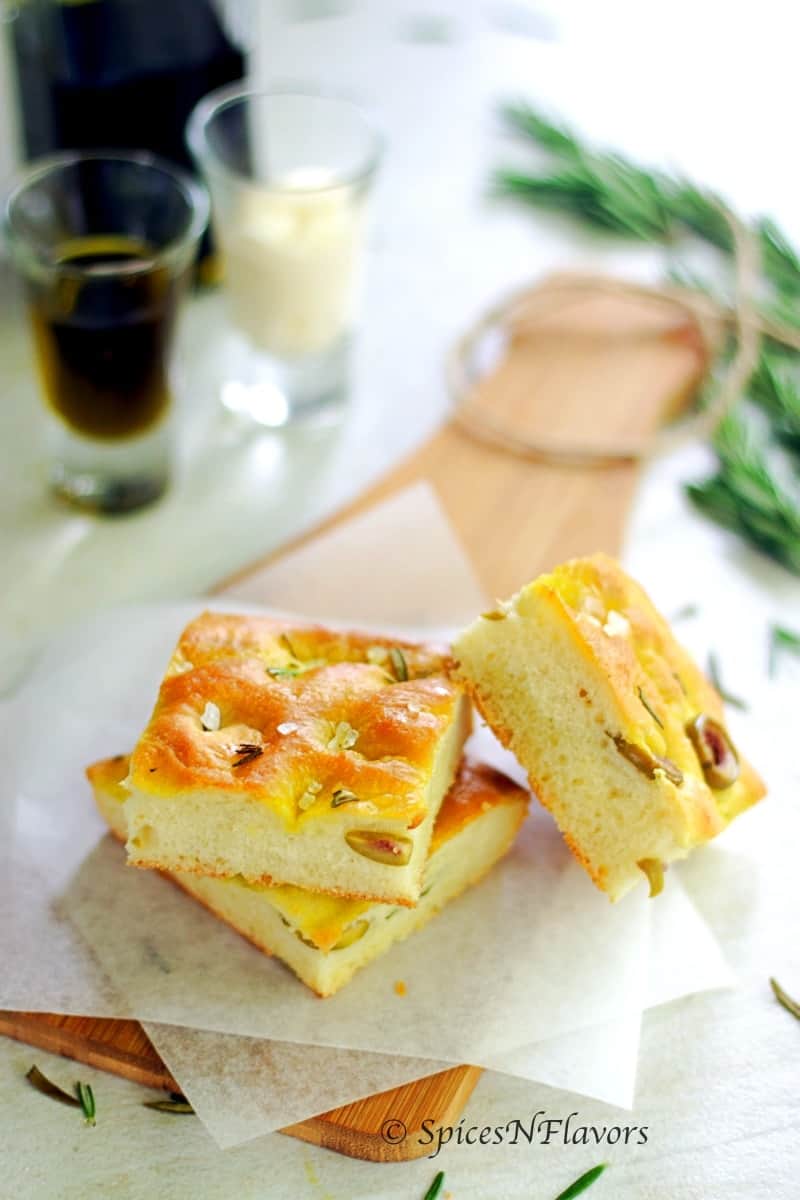 focaccia bread cut into squares and placed on a parchment paper on a wooden serving board with more olive oil and dipping sauce in the background
