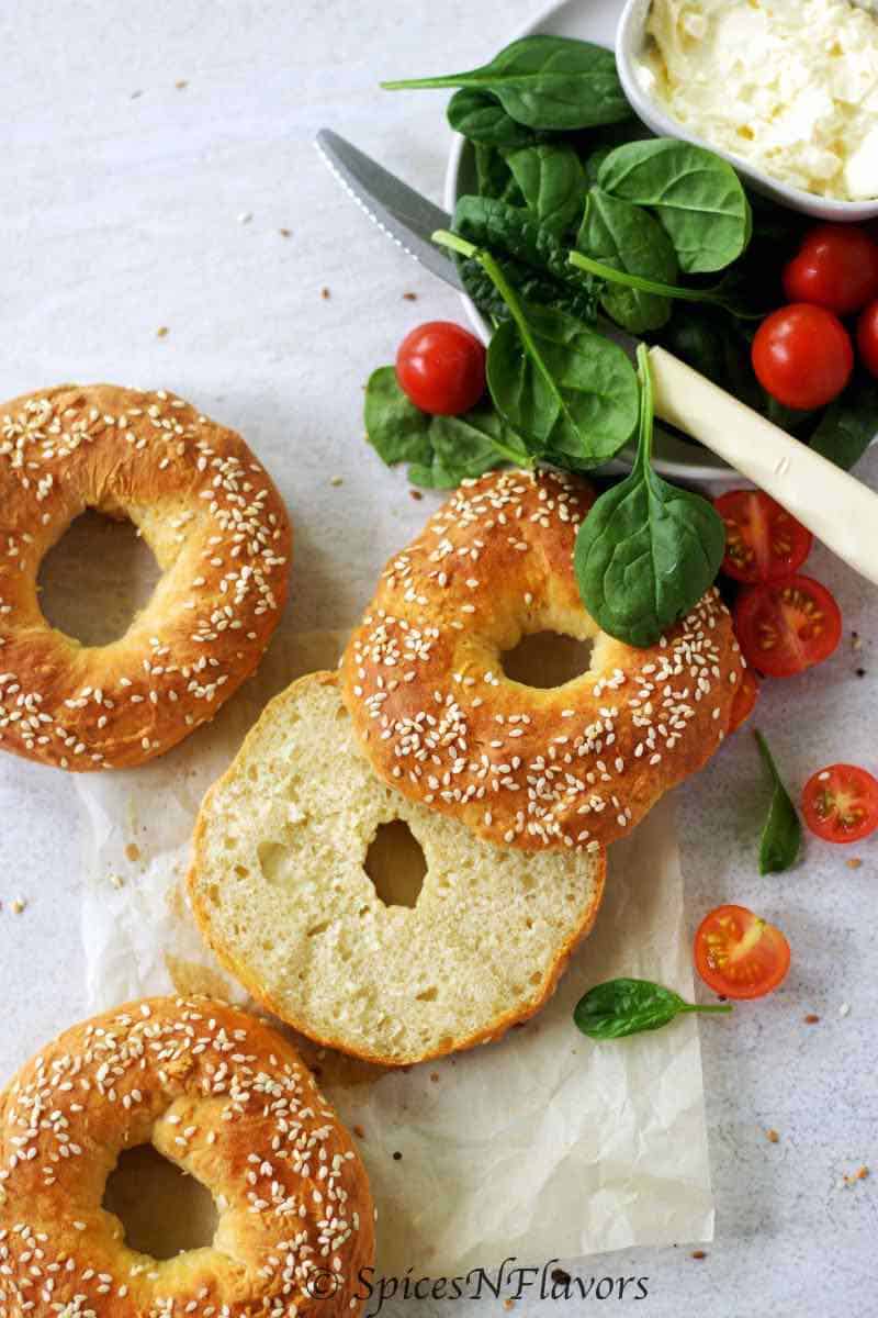 a bagel sliced horizontally and placed in the centre with 2 bagels on left sidr and some spinach and tomatoes on the right side