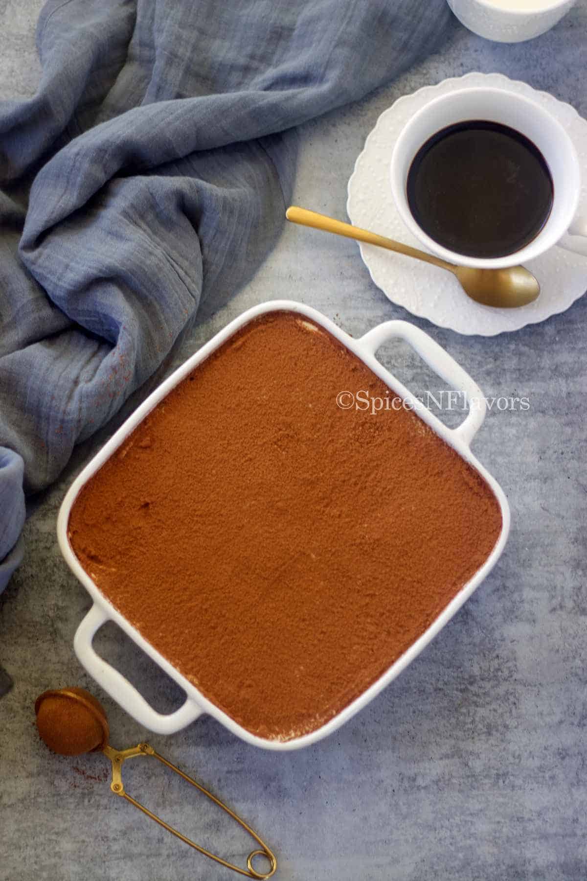 assembled tiramisu in a white baking dish dusted with cocoa powder served with black coffee on the side