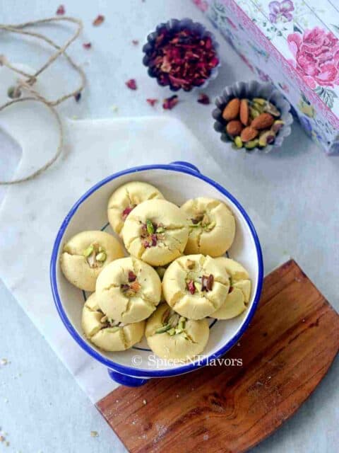 nankhatai biscuits placed in a wide mouth bowl over a marble cutting board with rose petals and nuts on the side