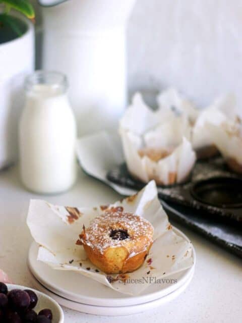 muffin piece placed on a white plate with milk, muffin tray and plant in the background
