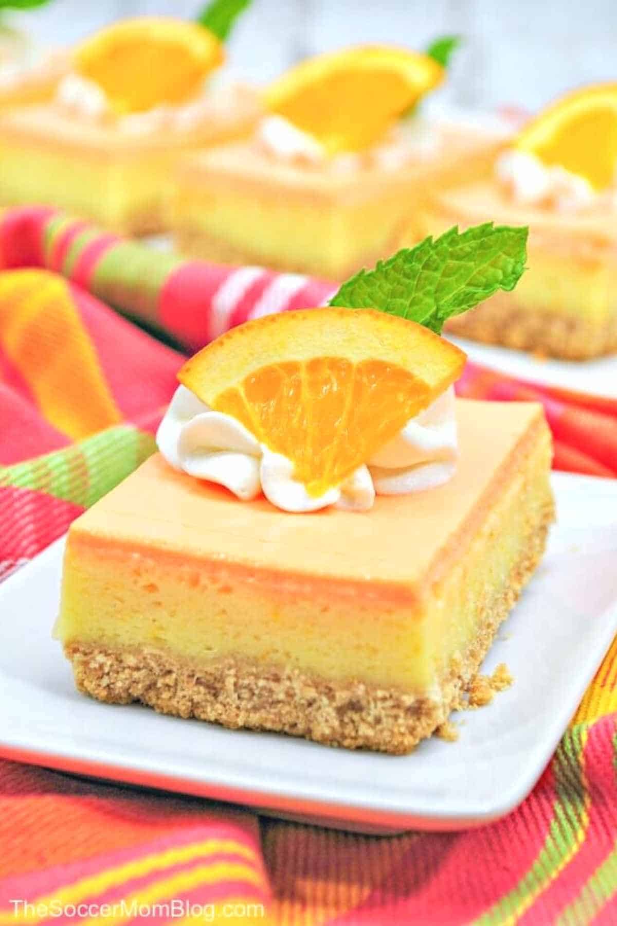 orange creamsicle cheesecake bars placed on a white plate with vibrant tea towel on the side