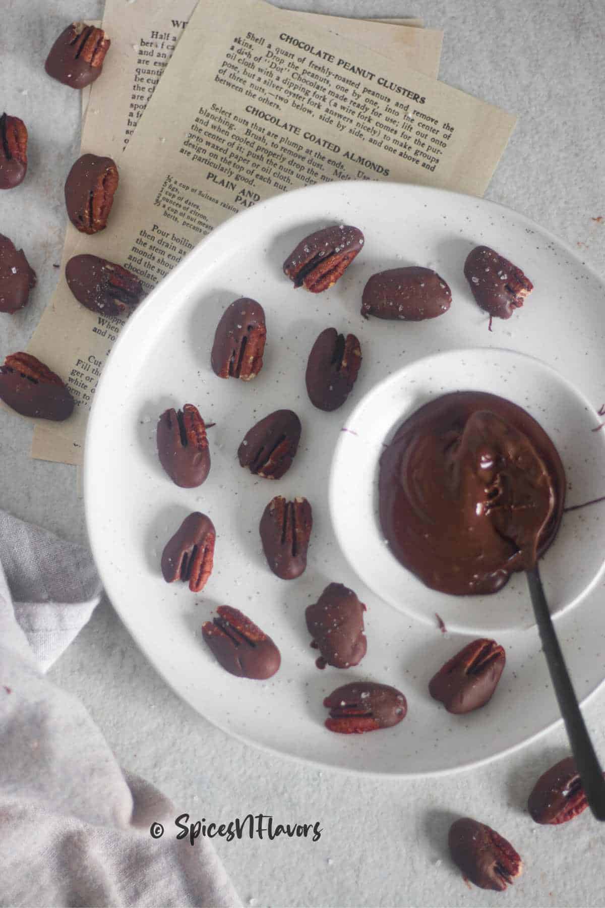 pecans covered in chocolate placed in a white round plate with a bowl of chocolate near to it