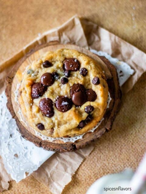 one chocolate chip cookie placed on a wooden coaster with melty chips on top